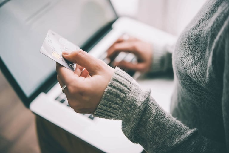 Uma mulher de blusa de lã cinza segurando um cartão de crédito na mão esquerda e mexendo no notebook que está apoiado em seu colo